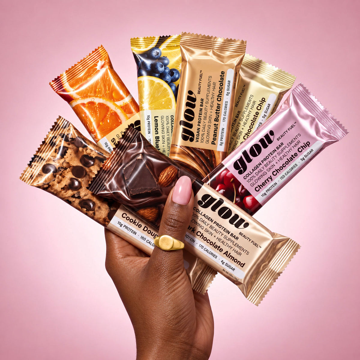 A hand holding a fan of glow protein bars in assorted flavors against a soft pink gradient background. The bars are individually wrapped in metallic foil with colorful fruit and chocolate graphics, including Orange, Lemon, Peanut Butter Chocolate, Blueberry, Cherry Chocolate Chip, Cookie Dough, and Chocolate Almond flavors.